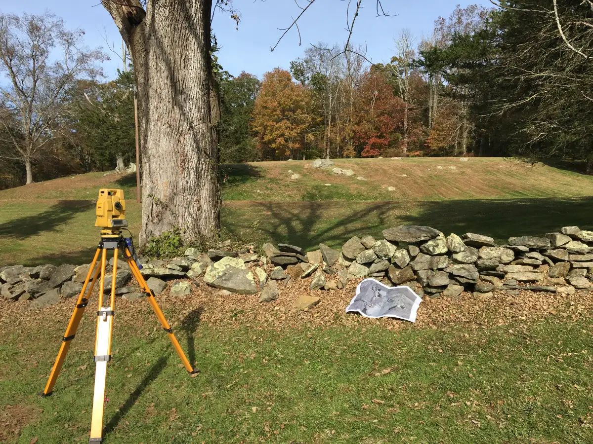 Total station on a tripod beside an old stone wall marking a property line